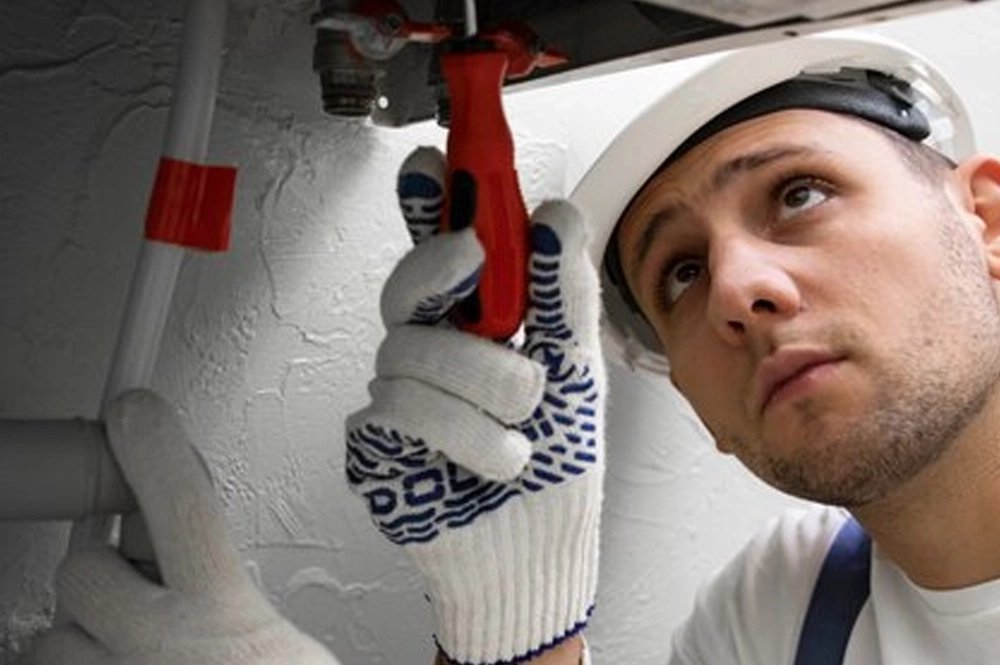 HVAC technician in a hard hat using a wrench to adjust a valve under a boiler