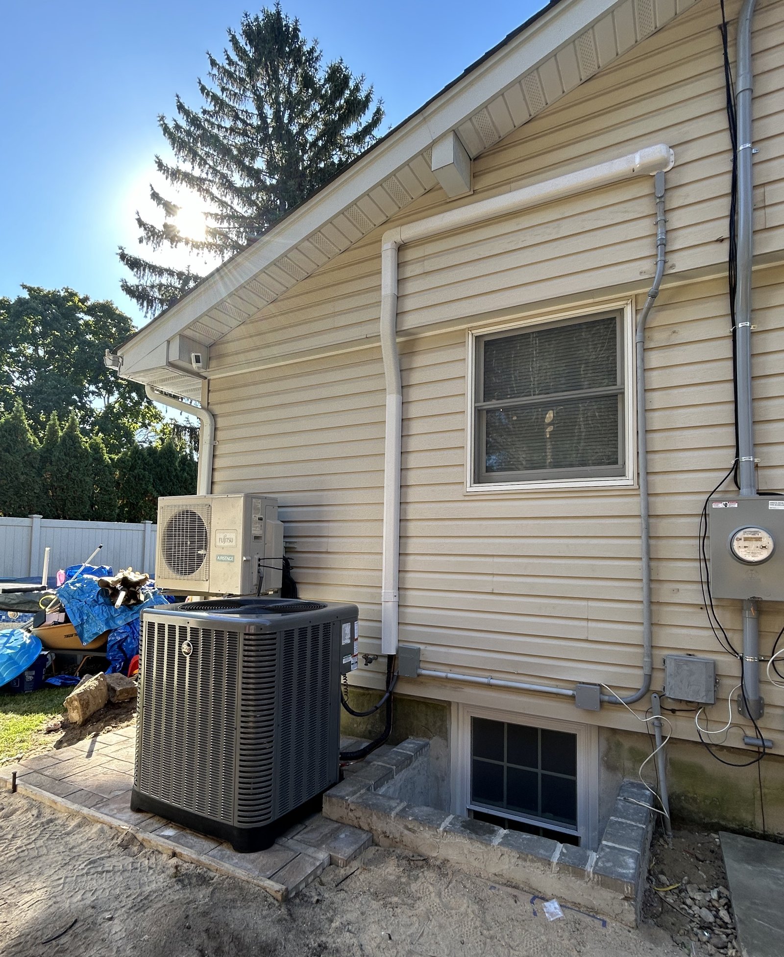 Outdoor installation of a Fujitsu mini-split unit alongside a central air conditioning condenser on the exterior of a house with beige vinyl siding